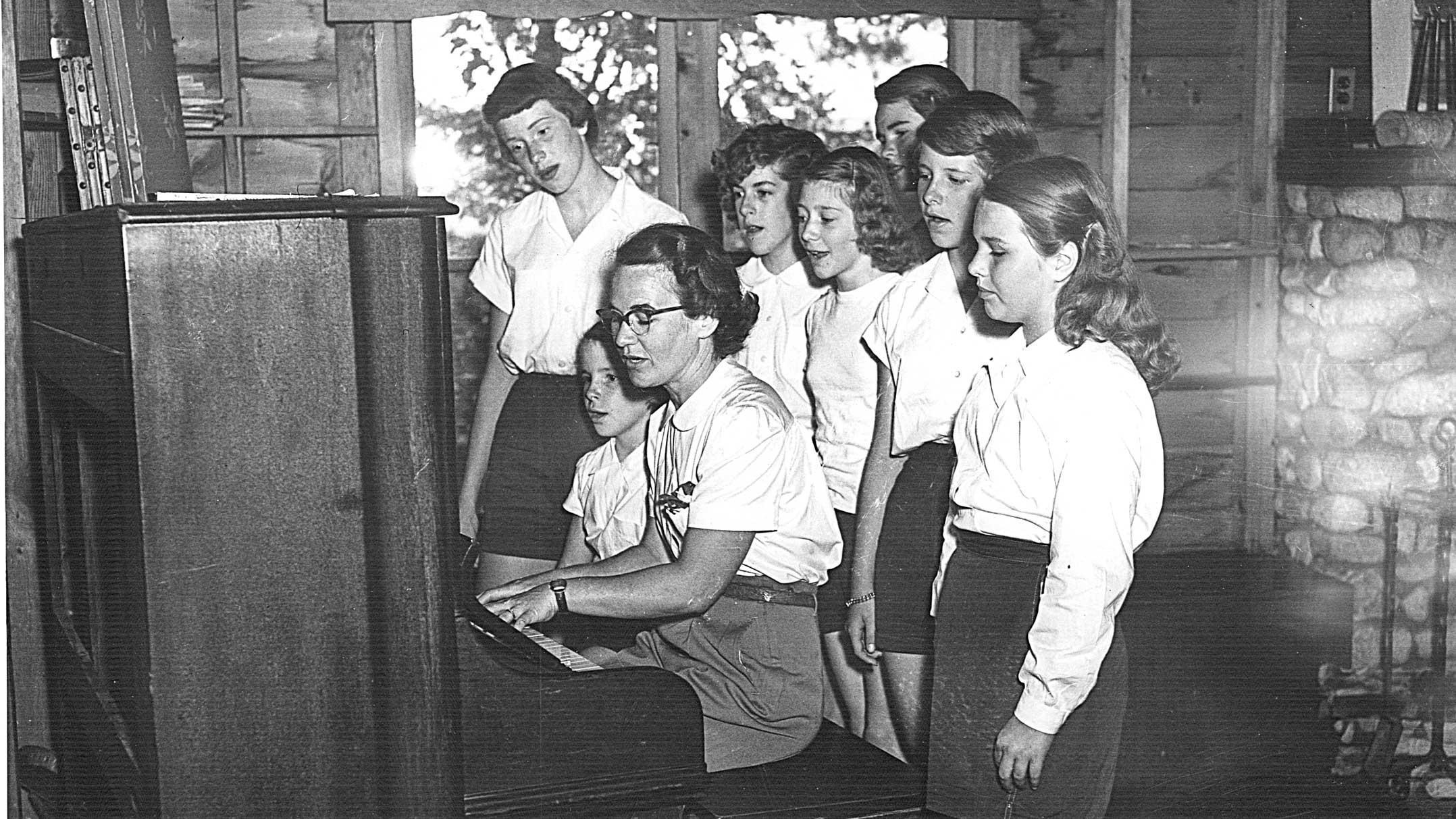 1951 photo of campers gathered around piano to sing