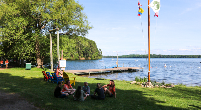 Campers sit in a circle near flagpole by lake