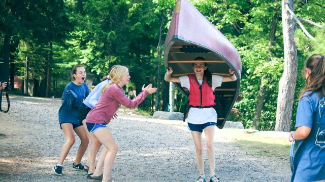 Camper carrying canoe while others cheer